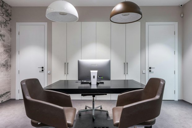 Armchairs at black table with modern computer placed under chandeliers in spacious stylish light apartment with white doors and closet
